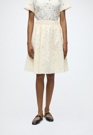 Naine kannab kreemikas silmkoega lühikeste varrukatega kleiti, millel on kogutud vöökoht, koos mustade Mary Jane tasaste jalanõudega.