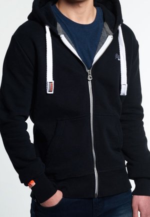 Personne portant un sweat à capuche noir zippé avec des cordons blancs sur un t-shirt bleu, mains dans les poches, visible du cou aux hanches.