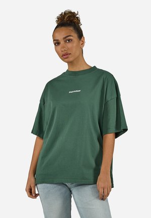 Kvinne med krøllete hår i en knute iført en løs mørkegrønn T-skjorte med hvit "PEGAADO"-tekst og lyseblå jeans, stående mot en ensfarget bakgrunn.