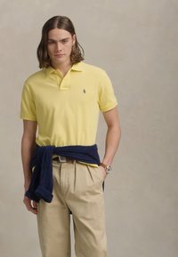 Tânăr cu părul până la umeri, îmbrăcat cu un henley galben, pantaloni bej, un pulover bleumarin legat în jurul taliei și un ceas de mână.