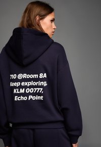 Tengerészkék kapucnis pulóver puha anyaggal, nagy fehér felirattal a hátoldalon: "710 @Room 8A Keep exploring, KLM 00777, Echo Point."