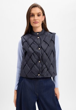 Frau trägt eine schwarze gesteppte ärmellose Weste mit silbernen Knöpfen über einem hellblauen Langarmhemd und dunkelblauer Jeans vor weißem Hintergrund.