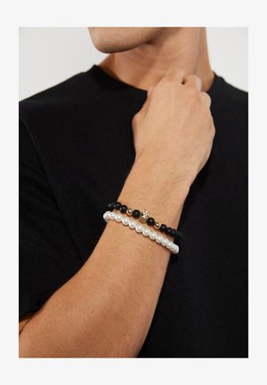 Conjunto de pulseras de cuentas que presenta perlas blancas y negras con cuentas mate negras, acentuadas por un detalle de calavera dorada. Llevadas en una muñeca.