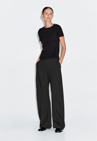 Schwarzes Strick-T-Shirt mit kurzen Ärmeln und Knopfdetails an der Schulter, kombiniert mit hoch taillierten, schwarzen weiten Hosen und glänzenden schwarzen Schuhen.