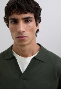 Suéter de punto verde oscuro con escote en V y cuello corto en punta, superpuesto a una camiseta blanca. Textura suave, diseño informal.