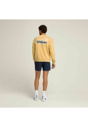 Mann trägt ein gelbes Wilson-Sweatshirt, marineblaue Shorts, weiße Socken und weiße Turnschuhe, steht mit dem Rücken zur Kamera vor einem einfarbigen Hintergrund.
