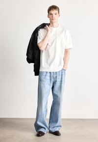 Tricou alb din bumbac cu decolteu rotund, asortat cu blugi largi, de culoare albastru deschis, și o jachetă bomber neagră aruncată peste umăr.