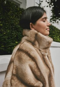 Faux fur coat in light beige with a high collar, featuring soft texture and subtle striped pattern. Worn by a person, side profile visible.