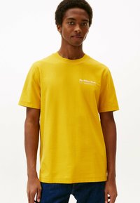 Junger Mann in einem gelben T-Shirt mit kleinen weißen Schriftzügen auf der Brust, der vor einem schlichten weißen Hintergrund steht.