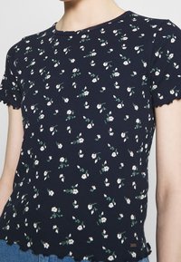 Top corto a maniche corte blu navy in tessuto a coste, con piccoli fiori bianchi e foglie verdi, orlo a onde e scollatura rotonda.