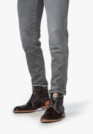 Botas de cuero marrón con acentos de ante texturizado, ojales de metal y una suela beige, combinadas con jeans de denim gris ajustados y doblados.