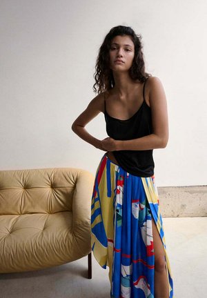 Giovane donna che sistema una gonna colorata con motivi blu, giallo e rosso con spacco sulla coscia, indossando un cami nero, in piedi vicino a un divano beige trapuntato.