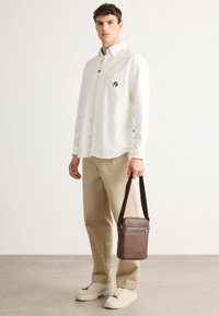 Jonge man in een wit overhemd, beige broek, crèmekleurige sneakers, met een bruine leren schoudertas met een ritsvak.