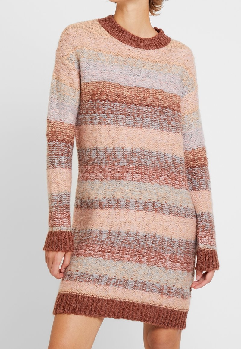 Femme portant une robe pull en tricot à manches longues avec des rayures horizontales dans des tons de rose, marron et gris clair.