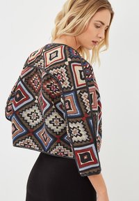 Chaqueta estampada con diseños geométricos en rojo, azul y beige. Confeccionada en un tejido texturizado con ribetes negros y mangas de tres cuartos.