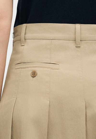 Κοντινή λήψη μπεζ πλισέ παντελονιών με θηλιές για ζώνη και κουμπωτή πίσω τσέπη, φορεμένα με μαύρο τοπ.