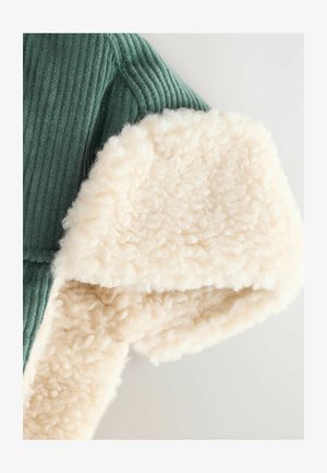 Il tessuto verde a coste incontra una morbida rifinitura in shearling crema, mostrando contrasti di texture e colori lungo il bordo di un indumento.