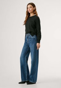 Pull en tricot noir, jean bleu à jambe large avec un motif subtil, porté avec des chaussures noires à bout pointu. Le modèle présente une tenue décontractée.
