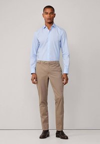 Camicia azzurro chiaro con bottoni e colletto, abbinata a pantaloni beige aderenti. L'outfit è completato da scarpe eleganti marrone scuro.