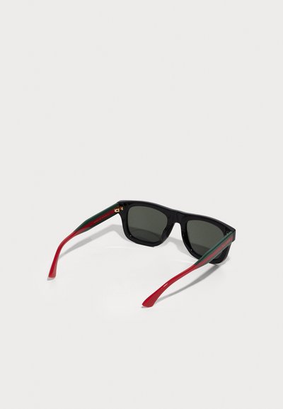 Óculos de sol pretos com lentes escuras, com braços listrados em vermelho e verde. A armação é retangular com uma textura suave e detalhes em dourado.