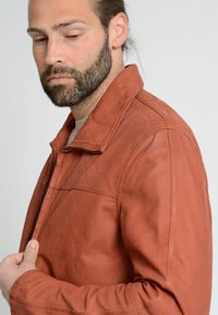 Braune Lederjacke mit Reißverschluss vorne, Stehkragen, horizontalen Nahtdetails über der Brust und einer glatten, leicht strukturierten Oberfläche.