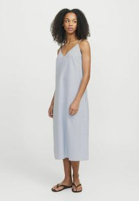 Vestido sin mangas a rayas azul claro con escote en V, silueta fluida, largo hasta la rodilla. Textura suave, bolsillos laterales, combinado con sandalias planas.