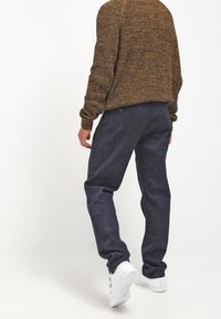 Person wearing a brown knit sweater, dark blue pants, and white sneakers, walking with back facing the viewer on a plain background.