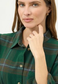 Camicia a quadretti verdi con strisce marroni e blu, colletto a bottoni, taschino sul petto e tessuto testurizzato. Modella che tocca il mento.