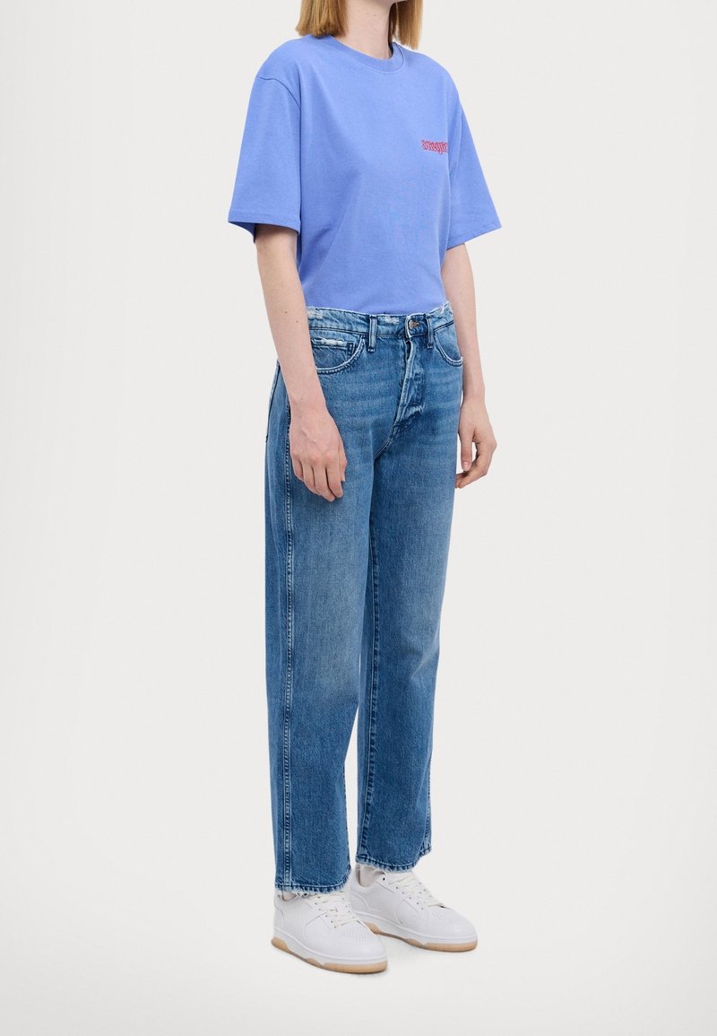 Vaaleansininen puuvillapaita, jossa on lyhyet hihat ja punainen logo; yhdistetty korkeavyötäröisiin vaaleansinisiin suoriin farkkuihin ja valkoisiin lenkkareihin.