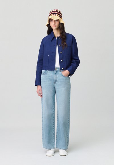 Veste marine courte avec boutons dorés, associée à un jean large bleu clair et des baskets blanches. Chapeau seau en crochet jaune avec des accents rouges et noirs.