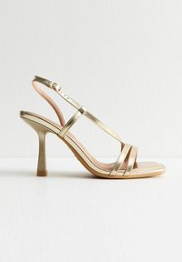 Guldmetalliska högklackade sandaler med tunna remmar, öppen tå-design och en smal stilettklack. Mjuk beige inredning.