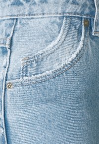 Tecido de denim azul claro com detalhes desgastados, um bolso frontal e um detalhe em botão prateado. Design clássico de cinco bolsos.