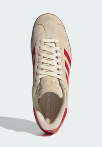 Μπεζ σουέτ αθλητικά παπούτσια με τρεις κόκκινες λωρίδες, λευκά κορδόνια, στρογγυλή μύτη και υφή στην εσωτερική σόλα που περιλαμβάν�ει το λογότυπο της Adidas.