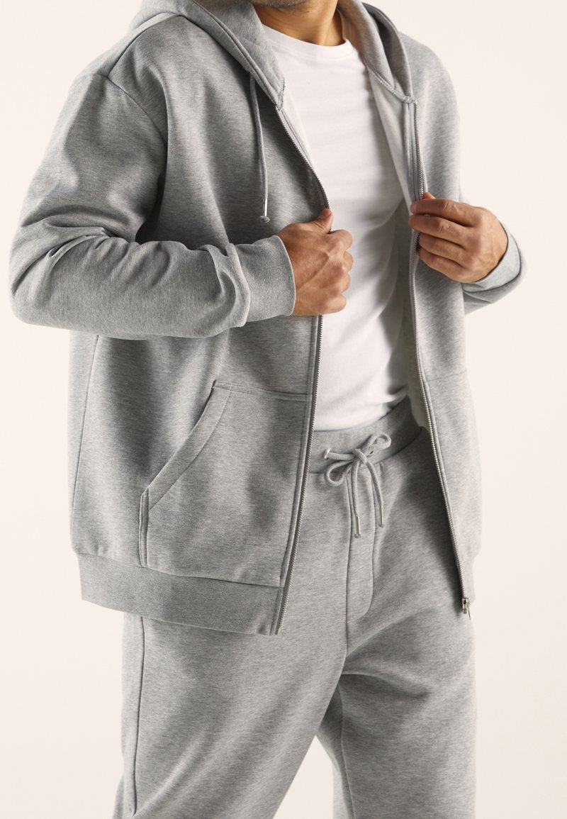 Homme portant un sweat à capuche zippé gris clair et un pantalon de survêtement assorti, ajustant la capuche sur un t-shirt blanc uni sur fond neutre.