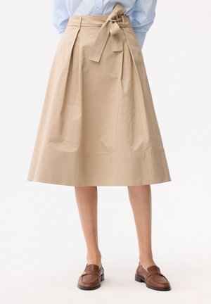 Béžová sukně střižená do A s širokým páskem uvázaným na mašli, nošená s světle modrou košilí a hnědými koženými mokasínami.