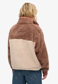 Brązowa i beżowa kurtka z polaru z wysokim kołnierzem, teksturowaną tkaniną i kontrastową dolną częścią. Posiada swobodny krój i elastyczne mankiety.