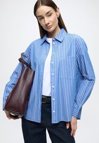 Cămașă cu dungi albastre și albe, cu nasturi și mâneci lungi, buzunar la piept, asortată cu o geantă burgundă texturată și blugi închiși la culoare.