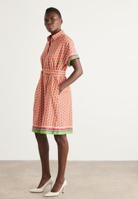 Kurzärmeliges Kleid mit einem leuchtend orangefarbenen Muster, das Früchten ähnelt, grünen Akzenten und einem Bindegürtel. Es verfügt über zwei seitliche Taschen und einen Kragen.