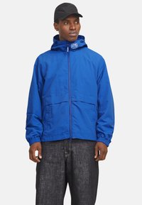 Leichte blaue Jacke mit Kapuze, Reißverschluss vorne und elastischen Bündchen. Verfügt über ein sichtbares Logo am Kragen und zwei Fronttaschen.