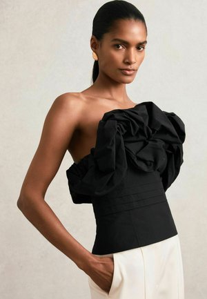 Femme portant un haut noir sans bretelles avec de grands volants et un pantalon blanc, posant avec une main dans la poche contre un fond neutre.