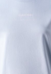 Světle modré bavlněné tričko s kulatým výstřihem, na přední straně s bílým potiskem "Calvin Klein" a "ZALOŽENO DEVATENÁCT ŠESTI OSMDESÁT".