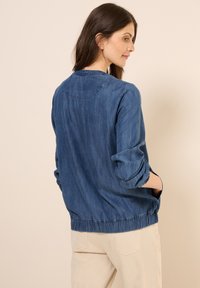 Jeansjacka i en medelblå färg med rund halsringning, elastisk midja och uppvikta ärmar. Har en avslappnad passform och mjuk textur.