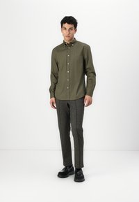 Camicia button-up verde oliva con colletto classico, abbinata a pantaloni grigi a motivo e scarpe nere, in piedi su uno sfondo bianco.