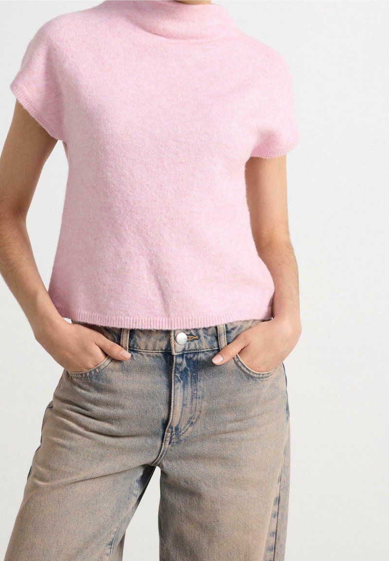 Femme portant un pull duveteux rose clair à manches courtes rentré dans un jean bleu délavé taille haute, les mains dans les poches.