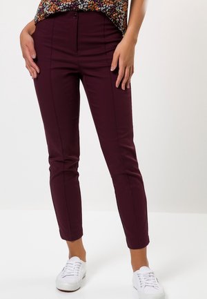 Frau trägt schlanke, knöchellange burgunderrote Hose und weiße Sneakers, mit einem teilweise sichtbaren, floral gemusterten Oberteil.