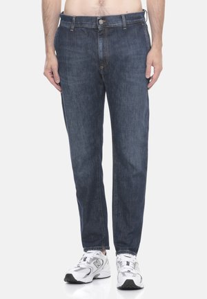 Uomo che indossa jeans blu scuro e sneakers bianche New Balance, mostrato dalla vita ai piedi su uno sfondo semplice.