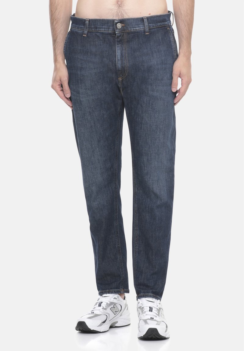 Uomo che indossa jeans blu scuro e sneakers bianche New Balance, mostrato dalla vita ai piedi su uno sfondo semplice.