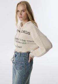 Haut en tricot beige coupé avec des accents de texte noir, associé à une jupe en jean bleu clair à taille haute et avec des poches.