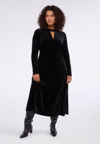 Černé sametové šaty s výstřihem ve tvaru klíče a dlouhými rukávy, pokryté bílými puntíky po celé látce. V kombinaci s vysokými černými kozačkami po kolena.