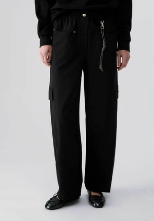 Persona che indossa pantaloni neri larghi con tasche laterali e un accessorio portachiavi, abbinati a scarpe nere e una maglia nera a maniche lunghe.